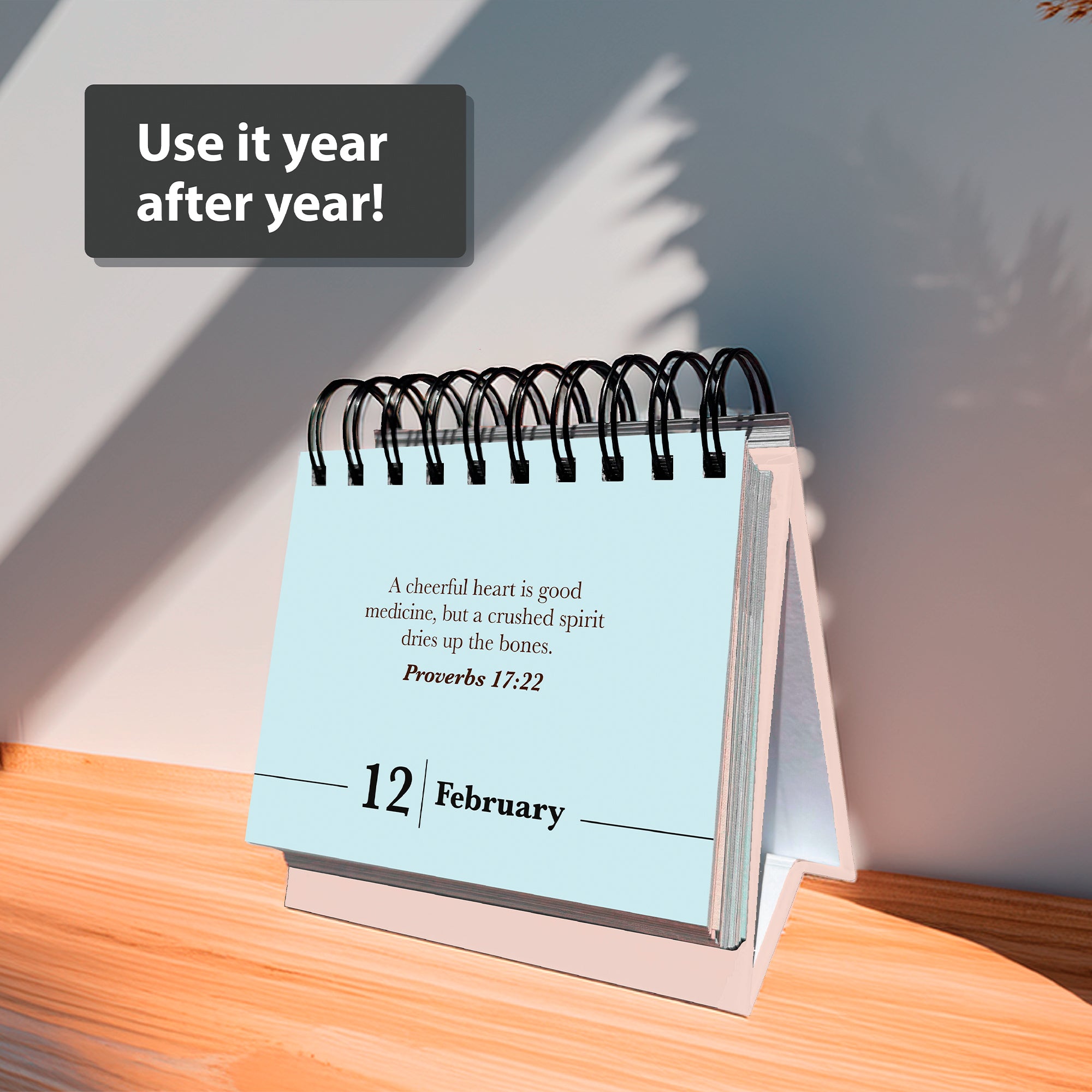 The HannahFaithstore Religious Perpetual Calendar displays a Bible verse for February 12. "Use it year after year!" appears above, as sunlight and a plant cast shadows on the wooden surface.