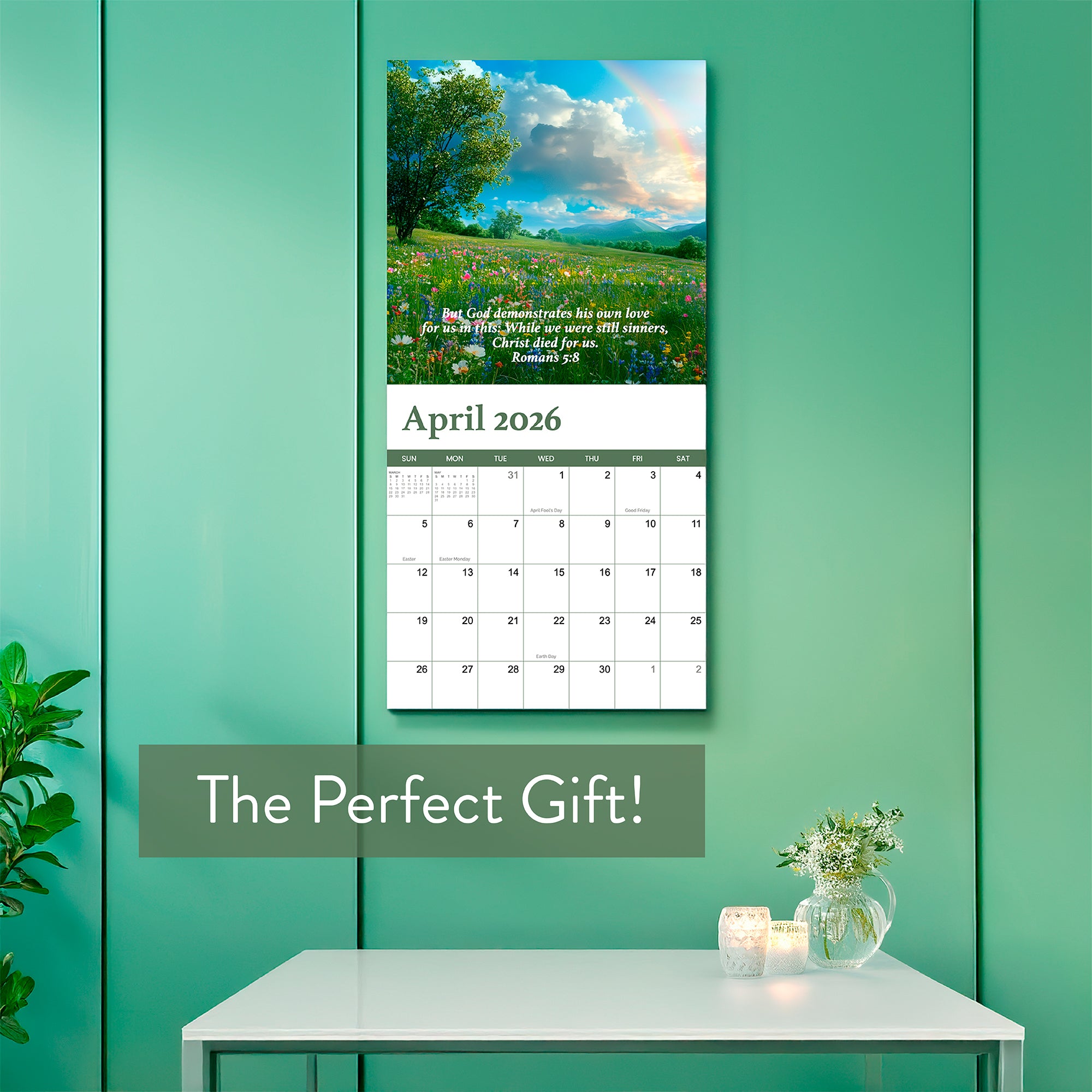 The 2026 Religious Wall Calendar by HannahFaithstore hangs open to April on a teal wall above a table with candles and a plant. It features a landscape photo, an inspiring Bible verse, and a rainbow—perfect as a thoughtful faith-based gift.