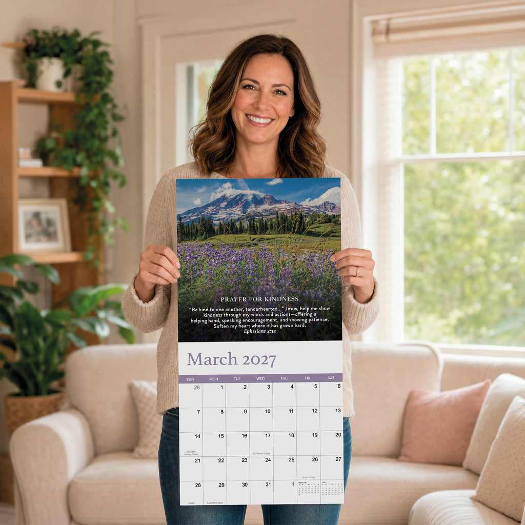 A woman stands in a living room holding the HannahFaithstore 2027 Prayer Wall Calendar open to March, displaying a stunning landscape photo and an uplifting prayer for kindness—a beautiful faith-based gift perfect for any home.
