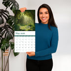 A woman in a blue sweater holds up a HannahFaithstore 2026 Religious Wall Calendar with a forest photo and Proverbs 3:6 above the dates—making it an ideal faith-based gift.