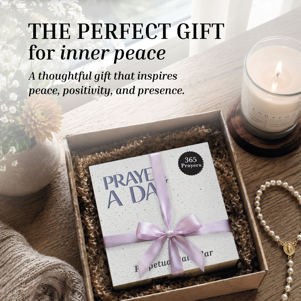 A boxed Perpetual Prayer A Day Calendar from HannahFaithstore, tied with a lavender ribbon, rests in a gift box beside a lit candle, flowers, and a cozy blanket—offering a thoughtful gift for peace and positivity.