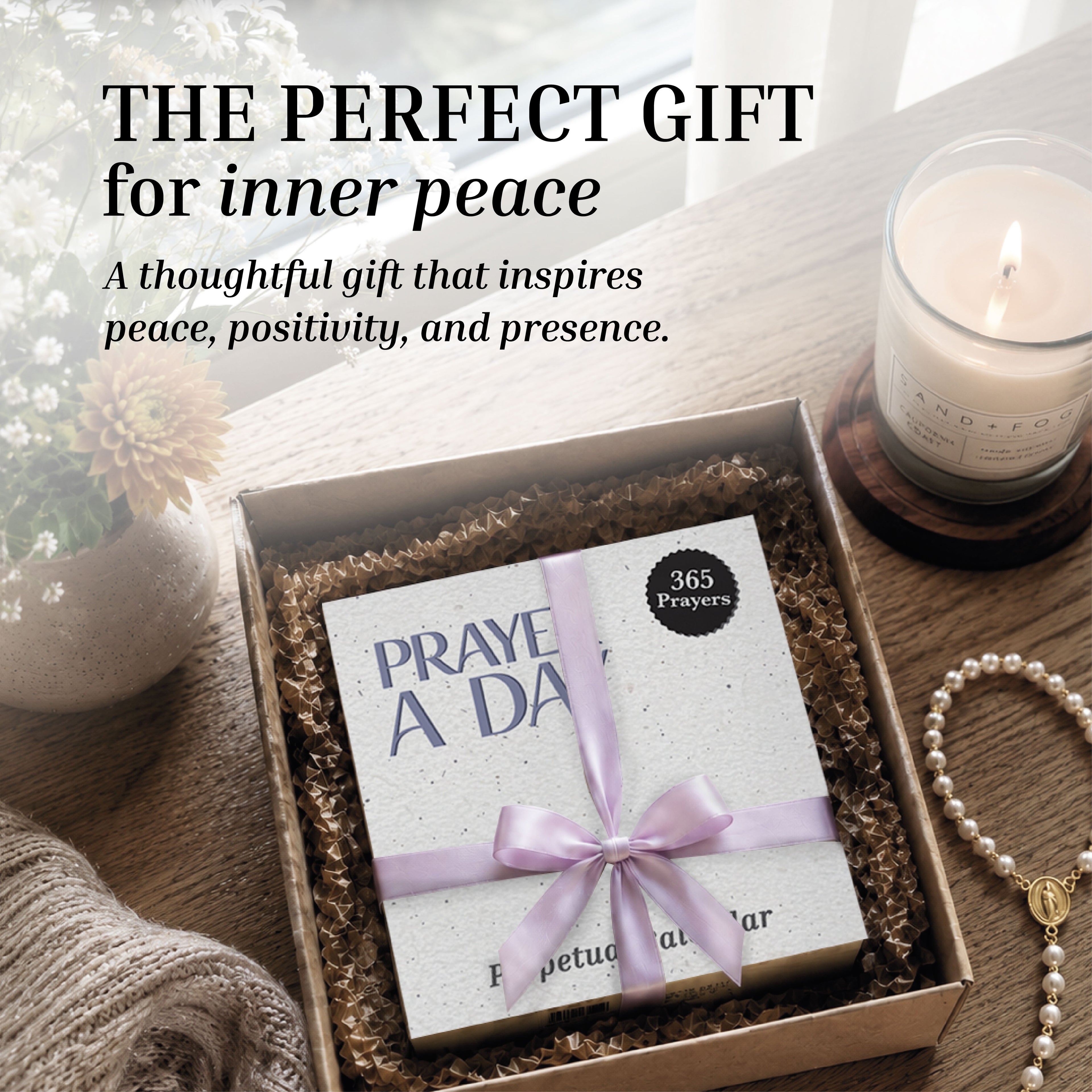 A boxed Perpetual Prayer A Day Calendar from HannahFaithstore, tied with a lavender ribbon, rests in a gift box beside a lit candle, flowers, and a cozy blanket—offering a thoughtful gift for peace and positivity.